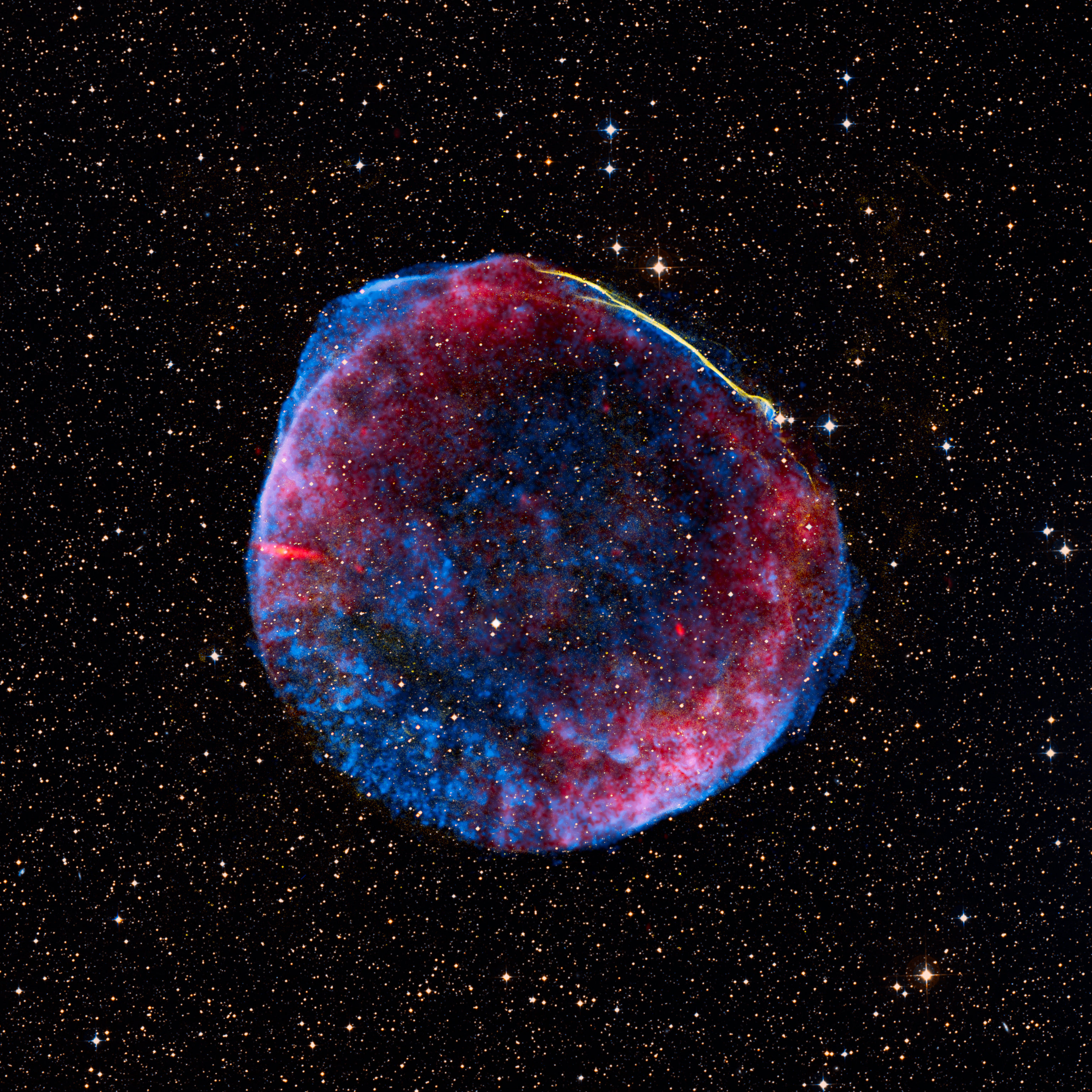 SN 1006: a composite image of a supernova remnant resembling a massive round red-white-and-blue firework against a densely starry sky. A colorful nebula with an electric blue hue and red and yellow highlights fills the center. Recent observations reveal the liberation of elements such as iron once locked inside the star.